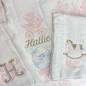 May include: White embroidered baby towels with pink and green designs. One towel features a pink baby carriage and the name "Hallie" in green. Another towel has a pink rocking horse and the name "Hallie" in pink. A third towel has the letter "H" in pink and green.