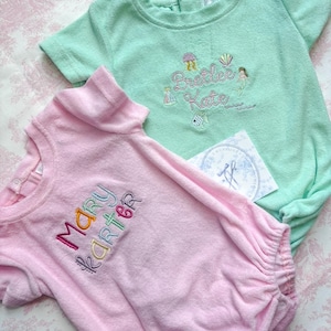 May include: Two terry cloth baby rompers, one light pink and one mint green. The pink romper has the name "Mary Karter" embroidered in colorful thread. The green romper has the name "Preetee Kate" embroidered with sea-themed designs.