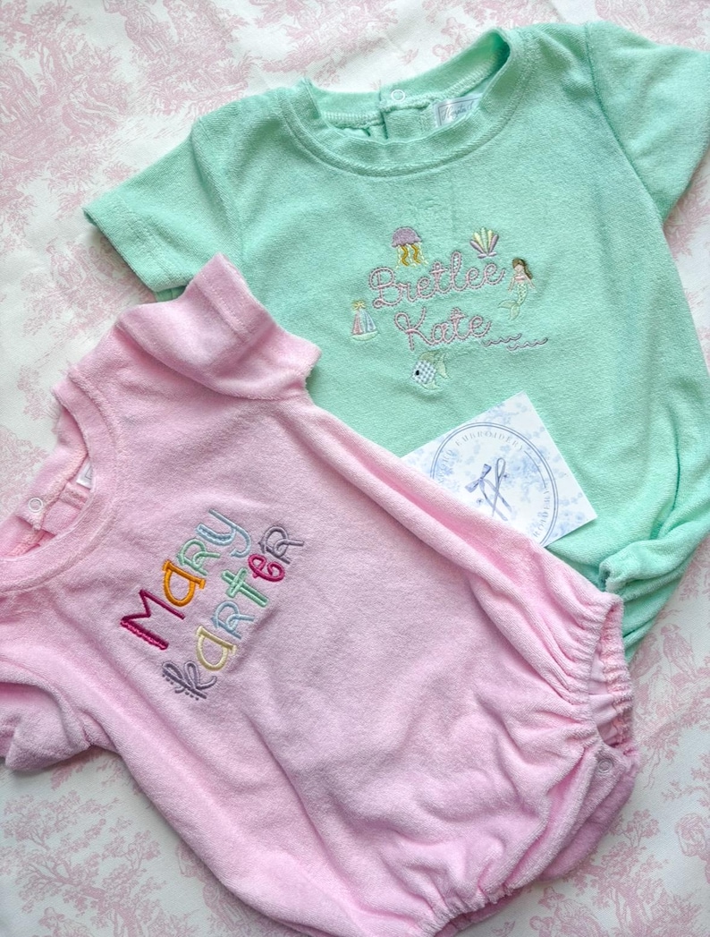May include: Two terry cloth baby rompers, one light pink and one mint green. The pink romper has the name "Mary Karter" embroidered in colorful thread. The green romper has the name "Preetee Kate" embroidered with sea-themed designs.