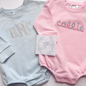 Może przedstawiać: Dwa pajacyki dla niemowląt, jeden jasnoniebieski i jeden różowy. Jasnoniebieski pajacyk ma wyhaftowane inicjały "HWC" w beżowo-biały wzór. Różowy pajacyk ma wyhaftowane imię "callie" we wzór kwiatowy. Oba mają długie rękawy.