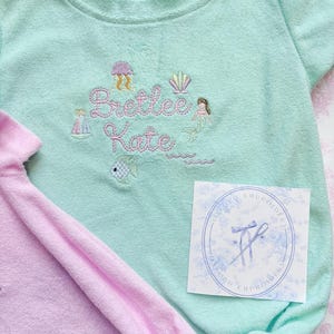 May include: A light teal baby romper with embroidered details, including a jellyfish, seashell, mermaid, sailboat, and the name "Bretlee Kate." A pink romper is partially visible. A white tag with a blue floral design is also present.