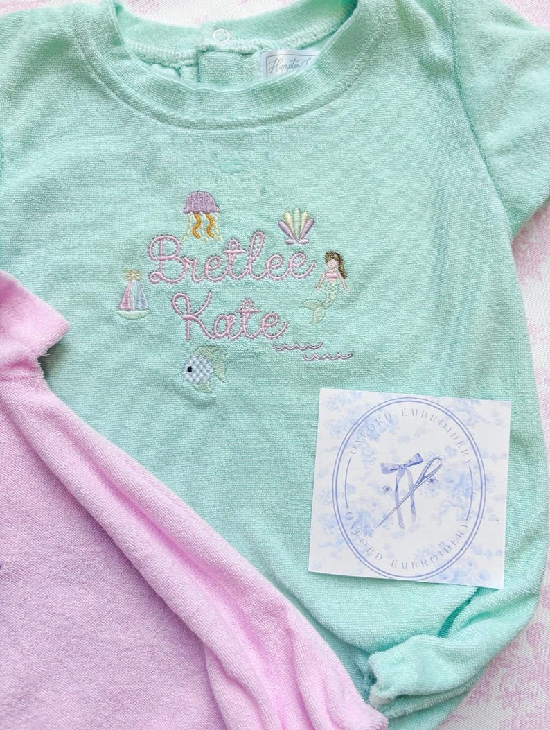 May include: A light teal baby romper with embroidered details, including a jellyfish, seashell, mermaid, sailboat, and the name "Bretlee Kate." A pink romper is partially visible. A white tag with a blue floral design is also present.