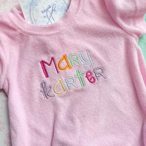 May include: A light pink terry cloth romper with short sleeves. The name "MARY KARTER" is embroidered in colorful thread across the chest. A tag with a blue floral design is visible near the neckline. The romper is designed for babies.