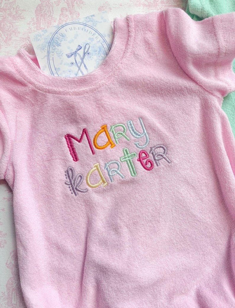 May include: A light pink terry cloth romper with short sleeves. The name "MARY KARTER" is embroidered in colorful thread across the chest. A tag with a blue floral design is visible near the neckline. The romper is designed for babies.