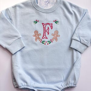 Op de afbeelding: Een lichtblauwe babyromper met lange mouwen. De romper heeft een geborduurd ontwerp van de letter "F" in roze, omlijst door een krans met peperkoekmannetjes en rode bessen. Het kledingstuk heeft drukknoopsluitingen aan de onderkant.