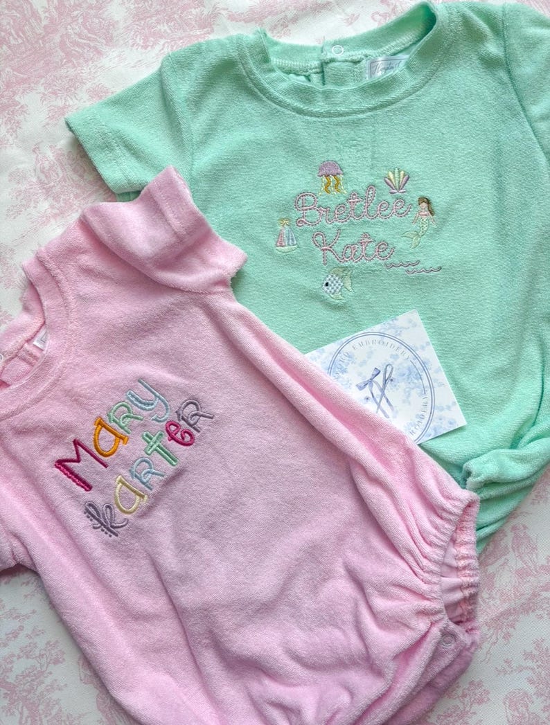 May include: Two baby rompers, one light pink and one mint green, made of soft terry cloth. The pink romper has the name "Mary Karter" embroidered in colorful thread. The green romper features the name "Brettee Kate" with mermaid and sea life embroidery.