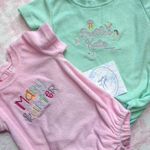 May include: Two baby rompers, one light pink and one mint green, made of soft terry cloth. The pink romper has the name "Mary Karter" embroidered in colorful thread. The green romper features the name "Brettee Kate" with mermaid and sea life embroidery.