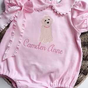 Pode incluir: Macacão rosa para bebê com gola branca e manga bufante. O macacão tem uma imagem bordada de um cachorro e o texto "Camden Anne".