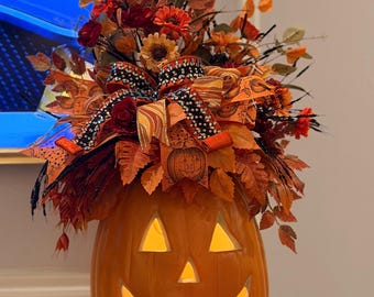 Puede incluir: Una calabaza de Halloween alta de color naranja con un rostro iluminado, rematada con un arreglo de flores artificiales de otoño, hojas naranjas y marrones, y una cinta decorativa con patrones de calabazas y detalles en negro brillante.