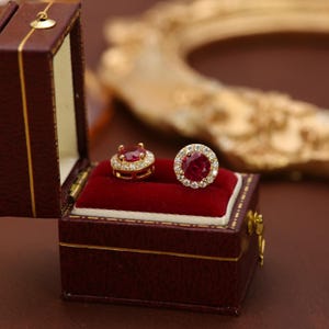 Puede incluir: Un par de pendientes de botón dorados con una gran gema roja central, rodeada de piedras claras más pequeñas. Los pendientes se exhiben en un joyero burdeos abierto con interior de terciopelo rojo.