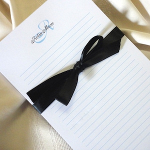 May include: A white notepad with blue lines and a black satin ribbon tied in a bow. The notepad has the name "Robin Moore" printed on the front in a blue and silver script font.