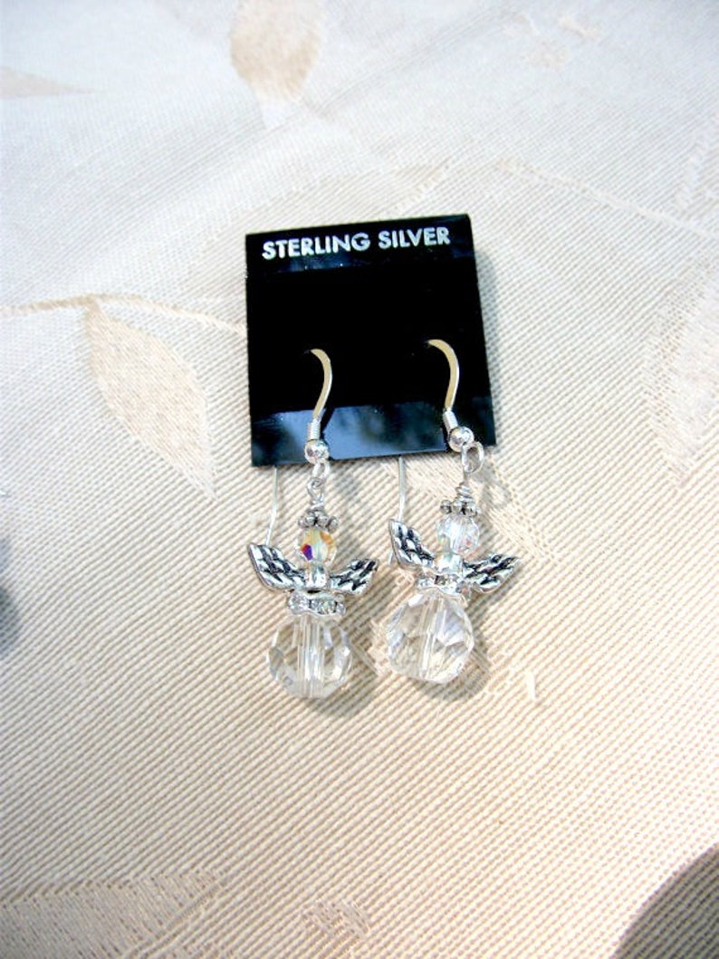 Puede incluir: Un par de pendientes de &aacute;ngel de plata de ley. Cada pendiente presenta un cuerpo de cristal transparente y facetado, alas plateadas y una peque&ntilde;a cabeza de cristal. Los pendientes se exhiben en una tarjeta negra con "STERLING SILVER".