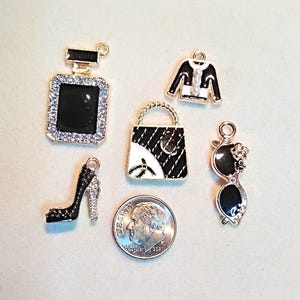 May include: A collection of five gold-toned metal charms featuring black and white enamel details, including a perfume bottle, a jacket, a handbag, a high-heeled shoe, and sunglasses, arranged next to a U.S. dime for scale reference.