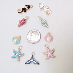 May include: Assortment of nine gold-tone metal charms with enamel detailing. The charms feature seahorse, seashell, starfish, and mermaid tail designs in various colours, including pink, blue, green, and black.