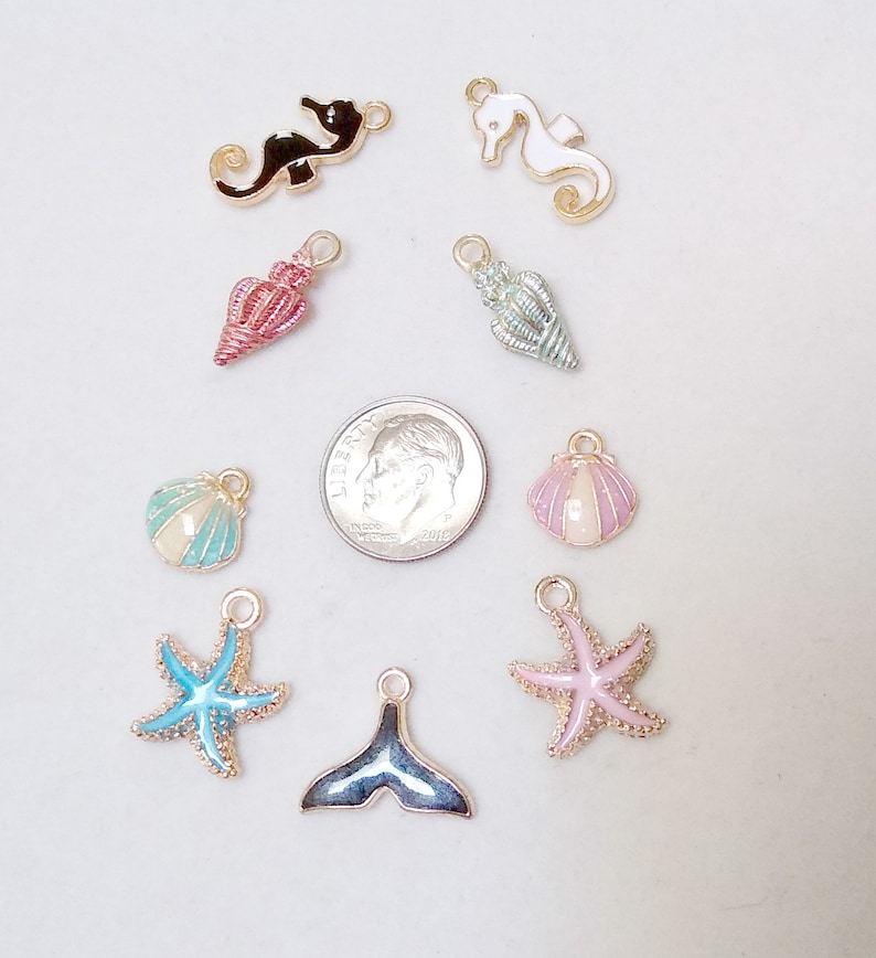 May include: Assortment of nine gold-tone metal charms with enamel detailing. The charms feature seahorse, seashell, starfish, and mermaid tail designs in various colours, including pink, blue, green, and black.