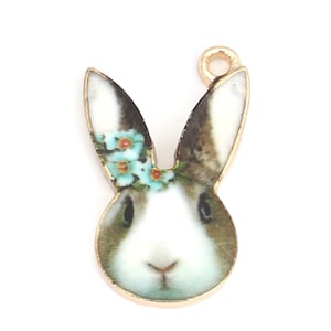 Könnte beinhalten: Goldfarbener Metallanhänger mit einem weißen Kaninchen mit braunen Ohren und einer blauen Blumenkrone.