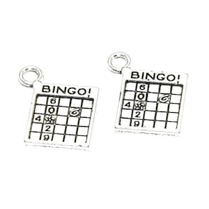 Puede incluir: Dos colgantes de plata con forma de tarjetas de bingo con la palabra "BINGO!" impresa en ellos. Cada tarjeta tiene los números 6, 20, 4, 2 y 9 impresos en ella.