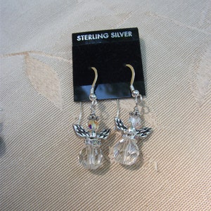 Puede incluir: Un par de pendientes de &aacute;ngel de plata de ley. Cada pendiente presenta un cuerpo de cristal transparente, alas plateadas y una peque&ntilde;a perla iridiscente. Los pendientes se exhiben en una tarjeta negra que dice "STERLING SILVER".