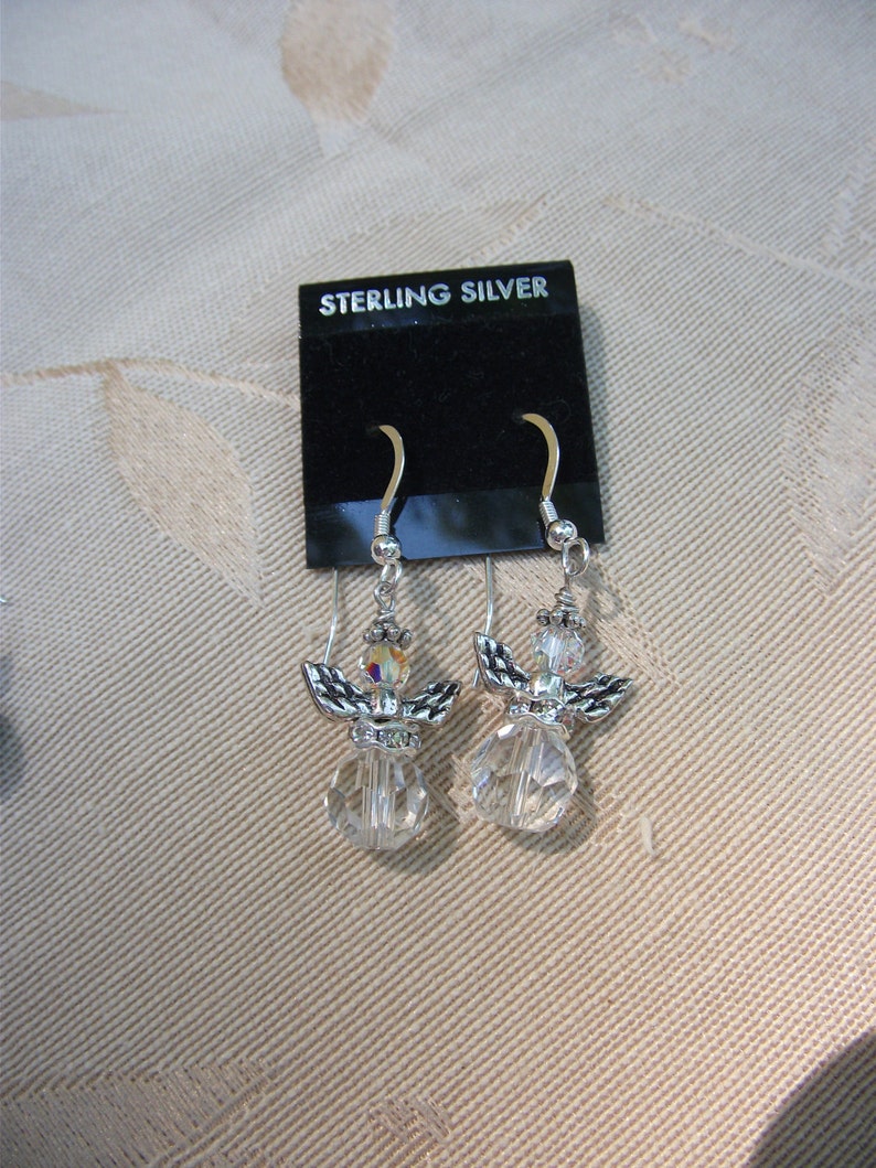 Puede incluir: Un par de pendientes de &aacute;ngel de plata de ley. Cada pendiente presenta un cuerpo de cristal transparente, alas plateadas y una peque&ntilde;a perla iridiscente. Los pendientes se exhiben en una tarjeta negra que dice "STERLING SILVER".