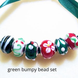 May include: A set of seven colorful glass beads strung on a green ribbon. The beads feature various designs, including stripes, swirls, and floral patterns in red, white, and green. The text "green bumpy bead set" is at the bottom.