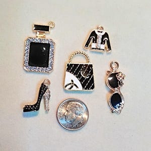 Puede incluir: Una colección de abalorios con temática de moda, que incluye un frasco de perfume negro con detalles de pedrería, un bolso blanco y negro, una chaqueta negra con una perla, un zapato de tacón alto negro y gafas de sol. Se incluye una moneda de diez centavos estadounidense como referencia de tamaño.