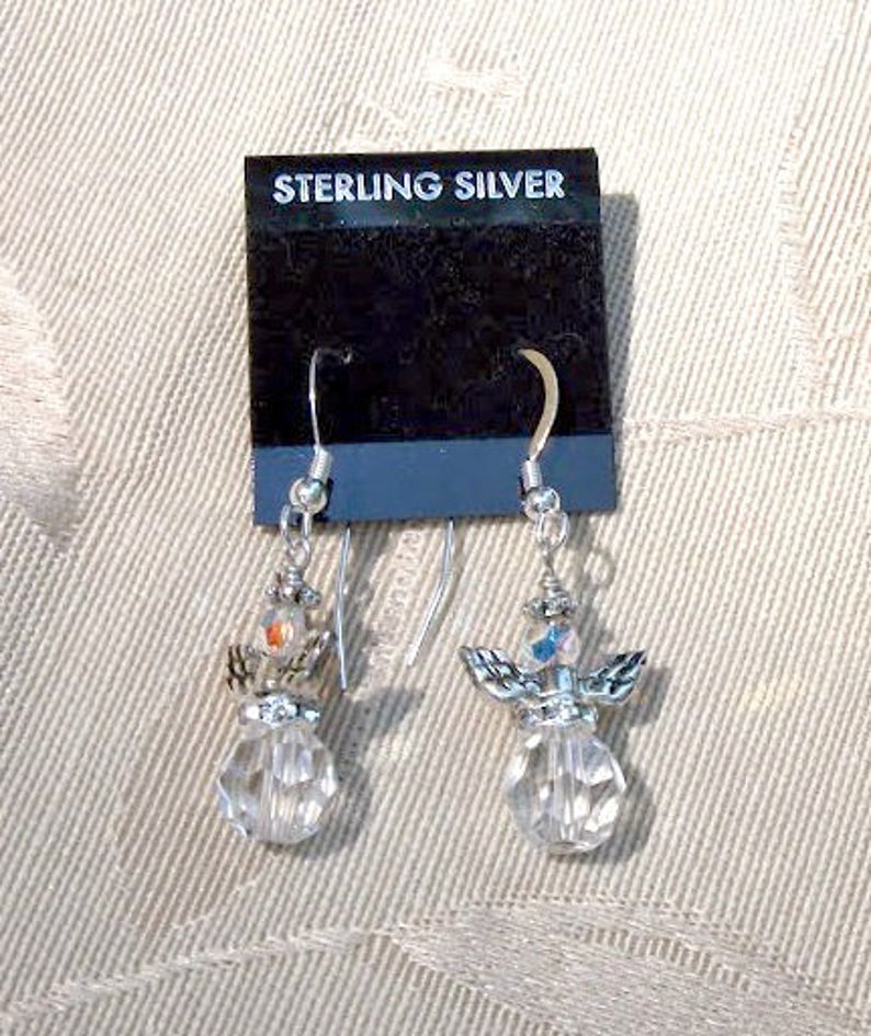 Puede incluir: Un par de pendientes de &aacute;ngel de plata de ley. Cada pendiente presenta un cuerpo de cristal transparente facetado, alas plateadas y una peque&ntilde;a cuenta colorida en la parte superior. Los pendientes cuelgan de ganchos plateados y se exhiben en una tarjeta negra con "STERLING SILVER".