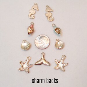 May include: A collection of gold and silver charm backs featuring seahorse, seashell, starfish, and mermaid tail designs. The charms are arranged on a white surface with a 10 pence coin for scale.