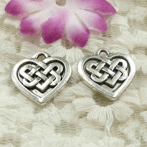 以下が含まれることがあります： ケルト結び目のデザインが施された銀色のハート型のチャームが2つ。チャームは白いレースの背景の上に置かれています。