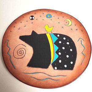 May include: Round, copper-colored decorative plate with a black bear silhouette. The bear has a patterned back with white dots and a yellow bird perched on top. The plate features celestial and geometric designs.