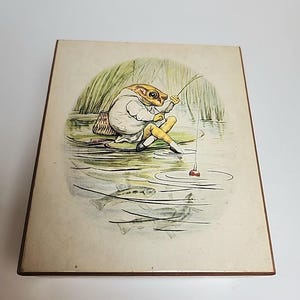May include: A vintage illustration of a frog wearing a white shirt and pants, sitting on a lily pad and fishing with a rod and line. The frog is surrounded by green reeds and the water is depicted with ripples and three fish swimming below the surface.