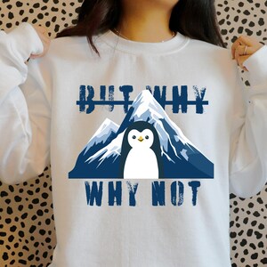 Könnte beinhalten: Weißes Sweatshirt mit einer Grafik eines Pinguins vor einer Bergkette. Der Text "BUT WHY" steht über dem Berg und "WHY NOT" darunter. Der Pinguin hat einen gelben Schnabel und ist mittig auf dem Sweatshirt platziert.