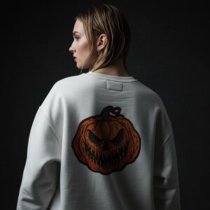 以下が含まれることがあります： 背中にハロウィーンのかぼちゃのデザインが刺繍された白いスウェットシャツ。 かぼちゃはオレンジ色で、恐ろしい顔をしています。 スウェットシャツはカジュアルなスタイルで、秋やハロウィーンに適しています。