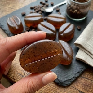 Op de afbeelding: Een doorschijnend, amberkleurig stuk zeep in de vorm van een grote koffieboon, met zichtbare donkerbruine spikkels over het hele oppervlak en een diepe centrale groef. De zeep heeft een gladde, gepolijste textuur en een afgeronde, ovale vorm.