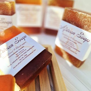 Op de afbeelding: Verschillende stukken African Soap worden getoond, sommige scherp, andere wazig op de achtergrond. De zeep is donkerbruin met een wit etiket. Het etiket leest "African Soap" en vermeldt ingrediënten. De zepen liggen op een houten zeepbakje.