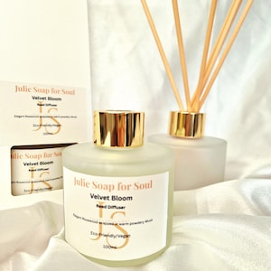 Puede incluir: Un difusor de varillas de vidrio esmerilado con una tapa dorada y varillas de madera. La etiqueta dice "Julie Soap for Soul Velvet Bloom Reed Diffuser". Una caja con la misma etiqueta está en el fondo. El producto es ecológico y vegano.