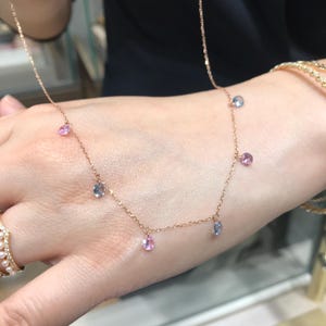 Può includere: Una delicata collana a catena d'oro con pendenti alternati di pietre preziose rosa e blu. La collana è sul polso di una persona.