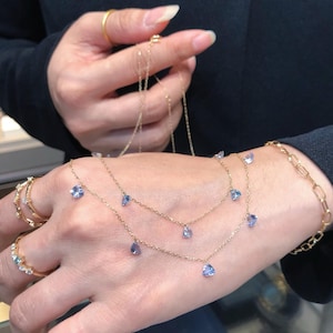 Puede incluir: Un delicado collar de oro con tres hilos, cada uno adornado con pequeñas piedras preciosas de color azul claro. El collar se muestra en una mano, mostrando el intrincado diseño. También son visibles varios anillos.