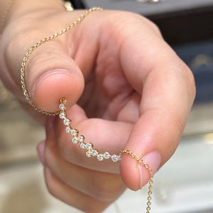 Puede incluir: Un collar de cadena dorada delicada con un colgante curvo engastado con un grupo de pequeñas piedras redondas, blancas y transparentes, dispuestas en un patrón escalonado de varios niveles que sigue un arco suave a lo largo de la cadena.