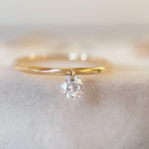 May include: A delicate gold ring with a single, sparkling round-cut diamond pendant. The ring band is thin and polished, and the diamond is suspended from a small loop. The ring is set against a soft, blurred background.