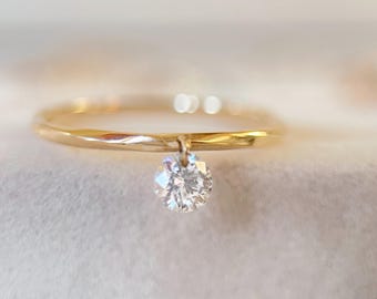 May include: A delicate gold ring with a single, sparkling round-cut diamond pendant. The ring band is thin and polished, and the diamond is suspended from a small loop. The ring is set against a soft, blurred background.