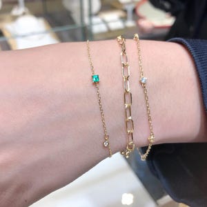 Op de afbeelding: Drie gouden kettingarmbanden met kleine edelstenen. De bovenste armband heeft een vierkante smaragdgroene steen, de middelste armband heeft een grotere, ronde, heldere steen en de onderste armband heeft een kleinere, ronde, heldere steen.