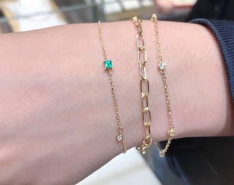 May include: Three gold chain bracelets with small gemstones. The top bracelet has a square emerald green stone, the middle bracelet has a larger, round, clear stone, and the bottom bracelet has a smaller, round, clear stone.