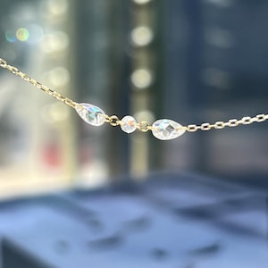 May include: A delicate gold necklace featuring three clear, faceted gemstones. Two teardrop-shaped stones flank a smaller, round stone. The chain is a fine link design, creating a minimalist and elegant aesthetic.