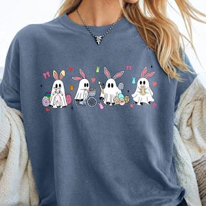 Puede incluir: Una camiseta azul con un diseño lúdico de cuatro figuras fantasmales vestidas como conejitos de Pascua. Cada conejito fantasma tiene diferentes accesorios, incluyendo huevos de Pascua y una bicicleta. La camiseta es parte de una colección de regalos hechos a mano.