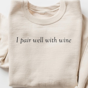 Könnte beinhalten: Ein beiger Pullover mit dem Aufdruck "I pair well with wine" auf der Vorderseite.