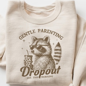 May include: A beige sweatshirt with a brown graphic of a raccoon wearing sunglasses and holding a coffee cup. The graphic says "Gentle Parenting Dropout Est. This Morning".