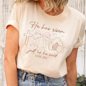 May include: Cream-colored t-shirt with the text "He has risen just as he said. Luke 24:6" and a graphic of a cross, bow, and tomb. The shirt is paired with light blue jeans.