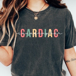 Peut inclure: Un t-shirt gris foncé avec le mot "CARDIAC" imprimé en lettres colorées. Les lettres sont soulignées de blanc. Le t-shirt est porté par une personne aux cheveux longs et bruns. La personne porte un jean bleu et des baskets blanches.