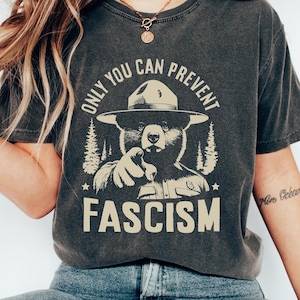May include: A dark gray t-shirt with a graphic of a bear wearing a ranger hat and pointing at the viewer. The text "Only you can prevent fascism" is printed on the shirt.