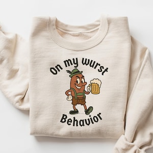 May include: A cream-coloured sweatshirt featuring the text "On my wurst behaviour" and a cartoon sausage character holding a beer stein. The sausage wears lederhosen and a green hat. Sleeves are rolled up.